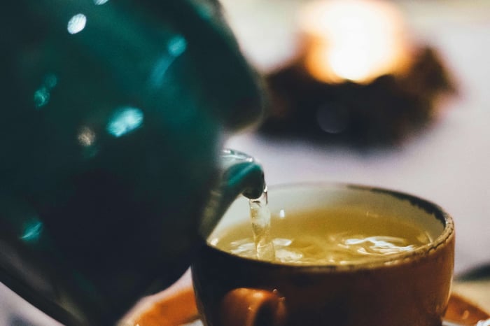 Green Tea Burn Fat is the question that underlies this scene of a green tea kettle slowly pouring a yellowish hued liquid into a tea cup.  The background is blurry and the emphasis is on the hot liquid being poured.